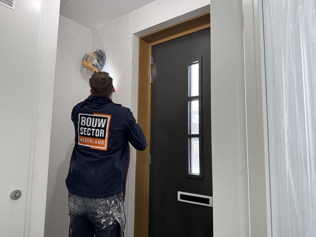 Vakman in een donkerblauwe Bouwsector-jas schuurt met een wandschuurmachine de wand naast de voordeur van een nieuwbouwwoning