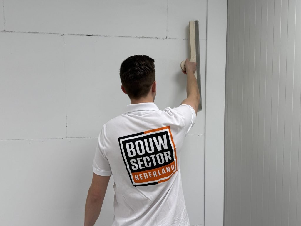 Stukadoor met het Bouwsector-logo op zijn rug houdt een spaan hoog tegen een wand van gipsblokken