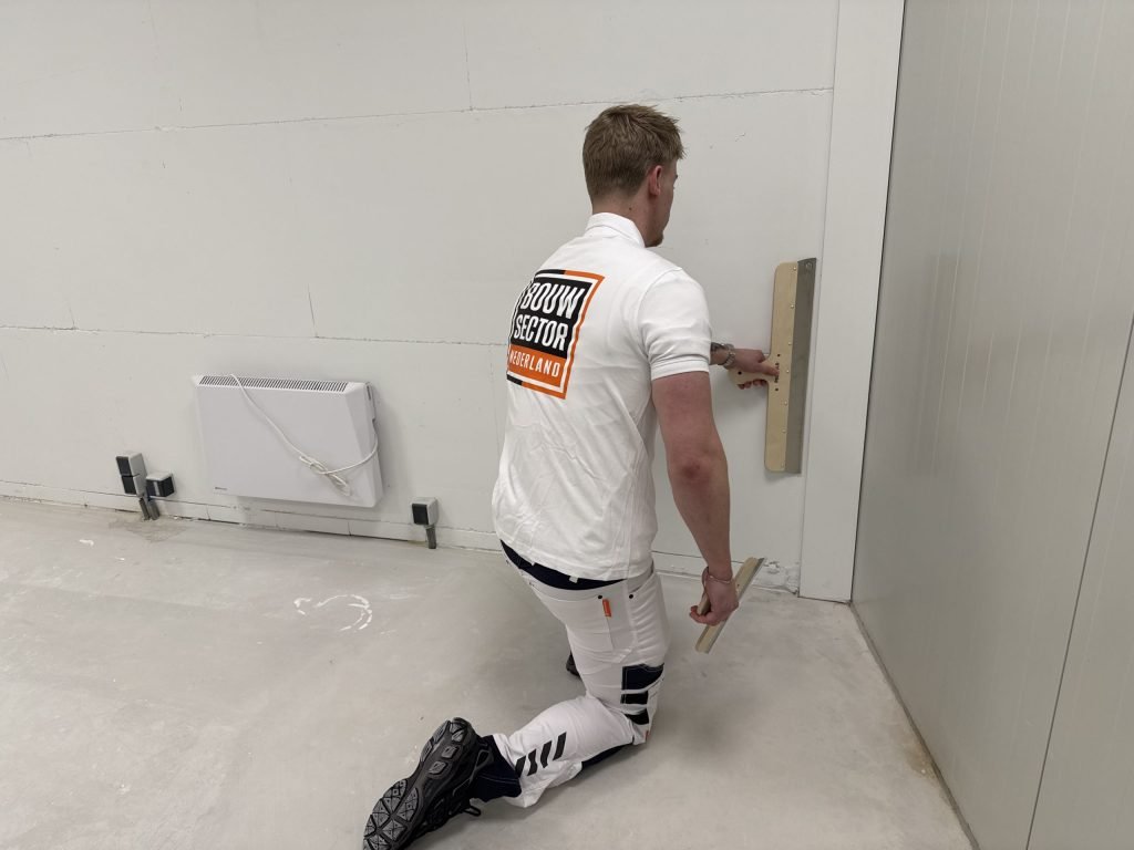 Stukadoor zit op een knie en strijkt met een brede spaan het onderste deel van een wand glad