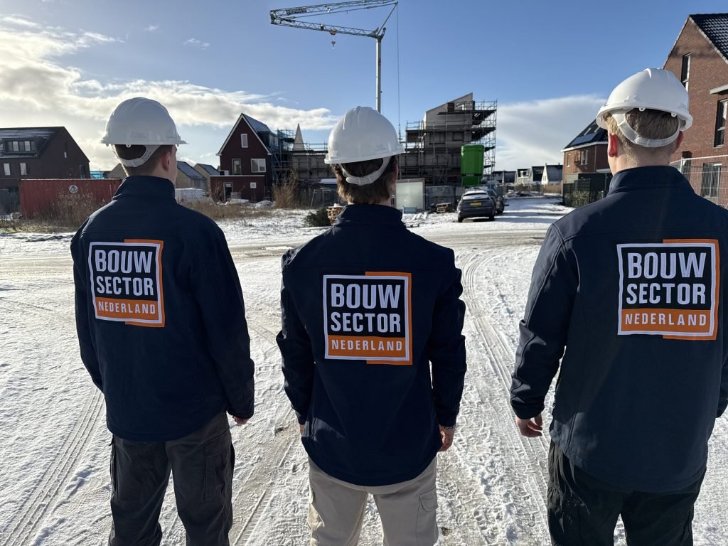 Drie vakmensen met witte bouwhelm kijken naar een rij woningen in aanbouw op een besneeuwde bouwplaats
