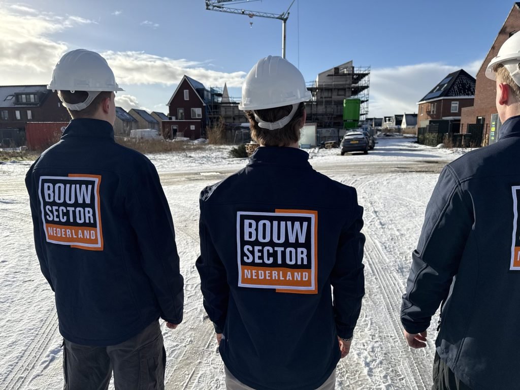 Drie vakmensen met witte helm en logo op de rug voor een rij huizen in aanbouw onder een blauwe lucht